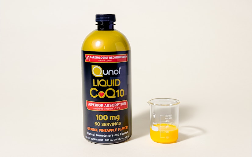 A bottle of Qunol liquid CoQ10 sits on a tan background with a small glass cup of the liquid next to it