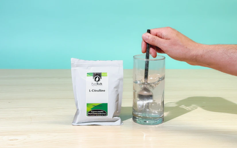 A pouch of PureBulk L-Citrulline next to a glass into which a hand is stirring the powdered supplement.