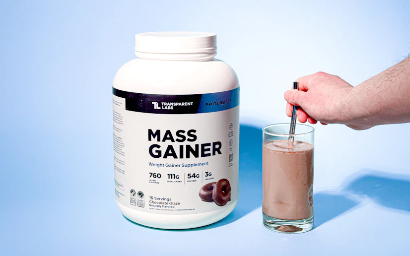 Transparent Labs Mass Gainer mixed in a glass next to its container against a blue background