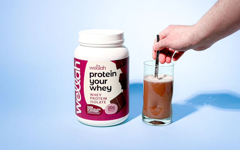 Wellah whey protein mixed in a glass next to its container against a blue background