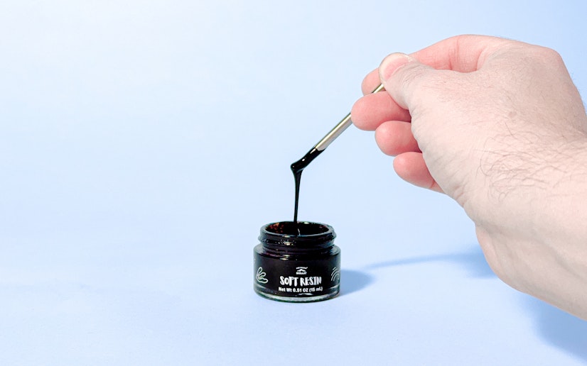 A hand holds a metal spoon pouring Pure Himalayan Soft Resin shilajit back into the jar.