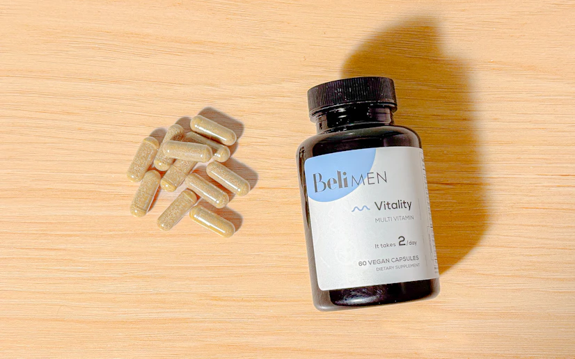 A bottle of Beli Vitality for Men with some of its capsules next to it laid out on a wood surface