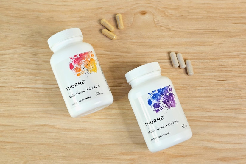 A top-down view of the Thorne Multi-Vitamin Elite A.M. and P.M. bottles on a wood countertop with three capsules of each supplement