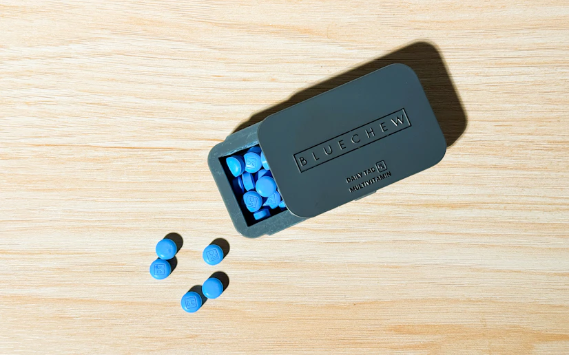 BlueChew daily tadalafil tablets spilling out of a gray tin on a wood surface.