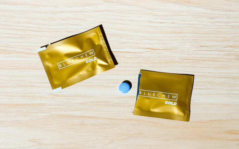 Two BlueChew Gold packets on a wood surface with one torn open and its tablet on the surface.