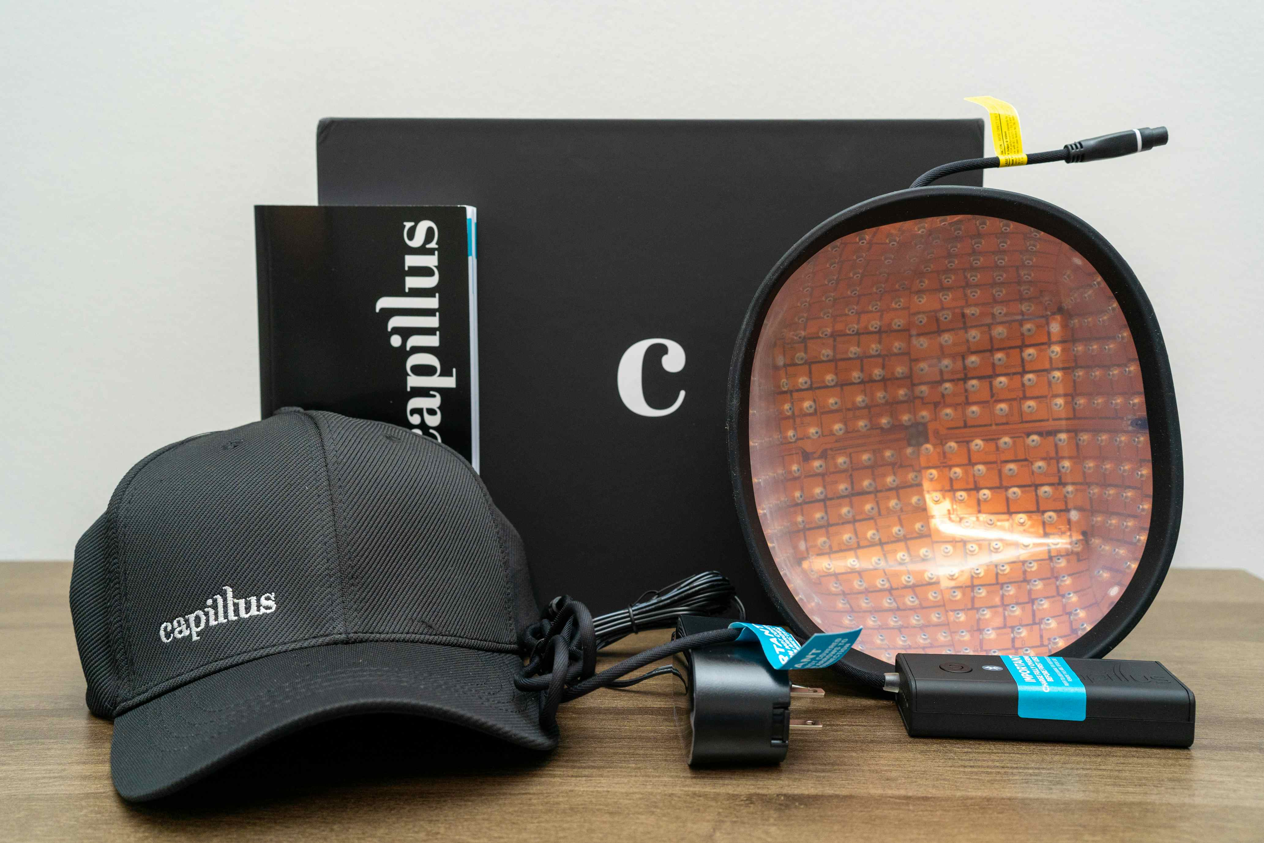 The image showcases a collection of items related to hair loss treatment, specifically featuring products from the brand capillus—a dark gray cap, a box, and a circular LED hair treatment device with wires and power supply.