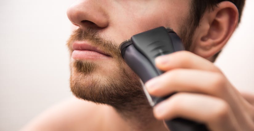Close-up of a man’s face in the mirror as he uses an electric razor to shave his light brown facial hair into a goatee as part of his morning hygiene ritual.