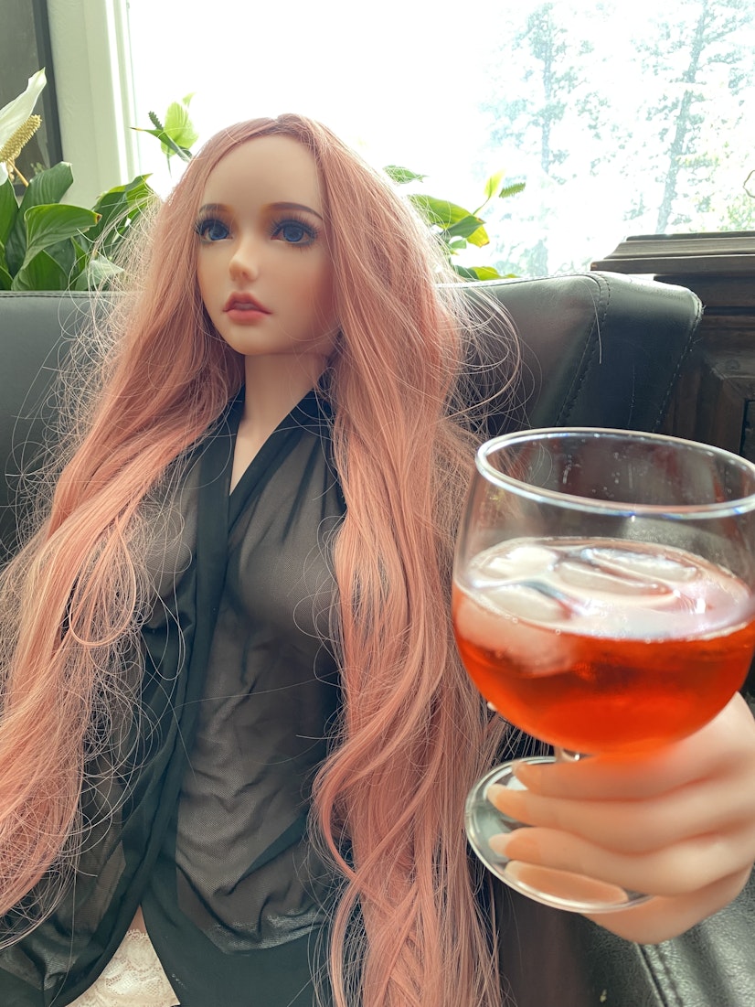 Realistic sex doll with long pink hair holding a glass of wine while seated on a couch