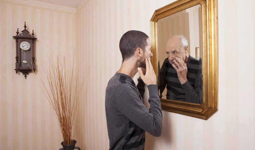A young man wearing a long-sleeved shirt with dark horizontal stripes looks into a mirror on the wall, holding his hands to his face, while his reflection shows an older, balding man with white hair and a wrinkled face looking back.